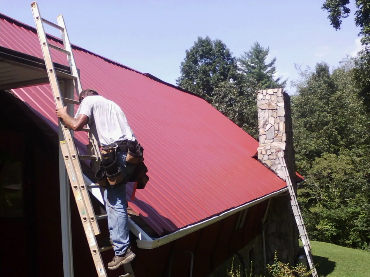 Roofer on a ladder beside a red metal roof and stone chimney — Soffit & Fascia Repair by Roofing Brothers in Steens